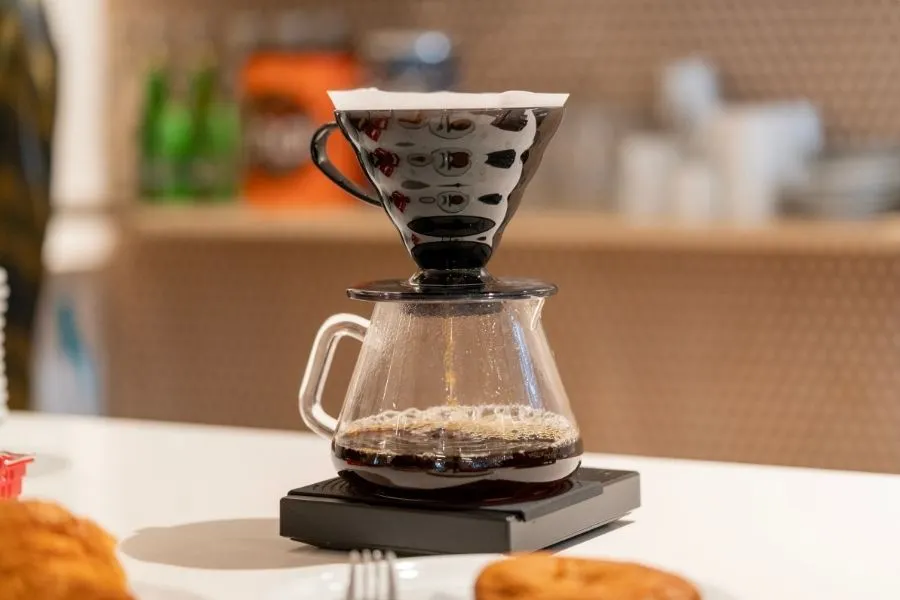 A pour-over carafe and V60 dripper on a wooden cutting board with freshly-ground coffee and a ceramic mug in soft morning light